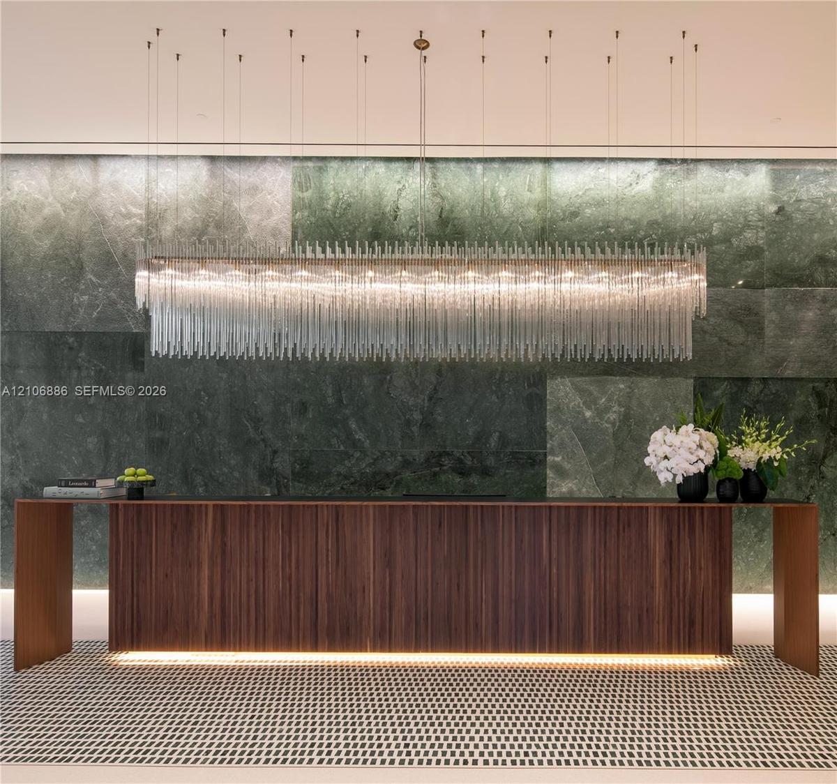 Large-scale linear chandelier with crystalline fringe, wood-veneer floating desk with base accent lighting, and full-height green-toned stone feature 