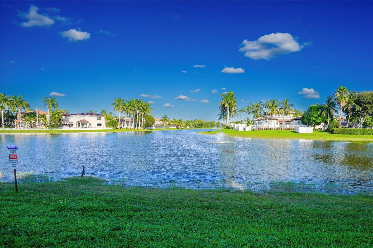 Waterfront property featuring a lake with a central fountain, manicured lawn, and mature palm trees