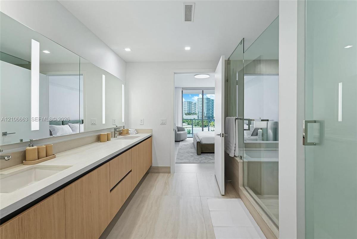 Dual-sink vanity with light wood-finish cabinetry, solid-surface countertop, full-width vanity mirror with integrated vertical lighting, and glass-enc