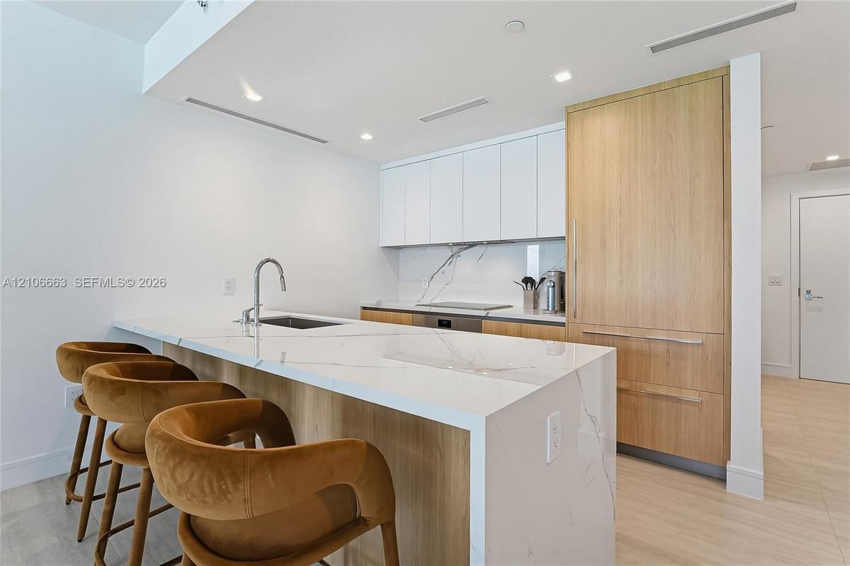 Kitchen featuring waterfall-edge stone countertops, custom cabinetry with wood-finish paneling, integrated refrigerator, stone backsplash, and stainle