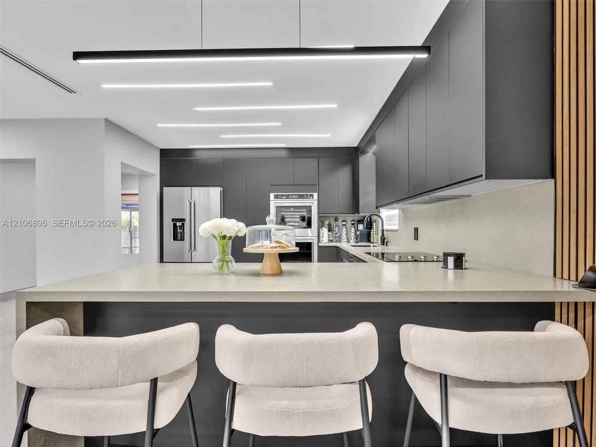 Modern kitchen featuring charcoal cabinetry, expansive stone-surface island, minimalist linear pendant lighting, built-in stainless steel double wall 