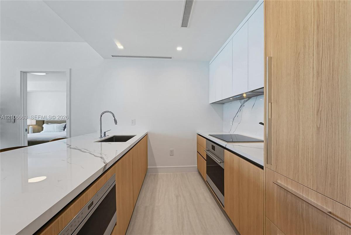 Contemporary kitchen featuring white flat-panel upper cabinetry, light wood-finish lower cabinetry, stone-style countertops with integrated backsplash