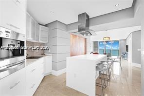 Modern kitchen featuring white flat-panel cabinetry, a central island with a solid-surface countertop, stainless steel vent hood, built-in coffee syst
