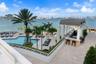 Waterfront pool terrace featuring a paved sun deck, built-in outdoor bar with a covered structure, mature palm trees, and perimeter landscape hedging