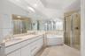 Double vanity with light-toned stone countertop, expansive wall-to-wall mirrors, recessed lighting, corner soaking tub, and enclosed shower with stone