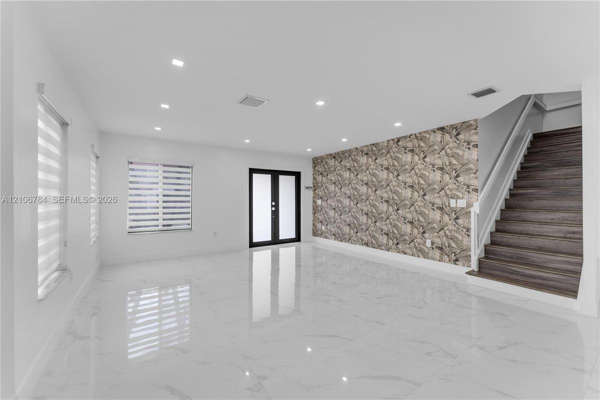 Open-concept room featuring large-format tile flooring, recessed lighting, double-door entryway, patterned accent wall, and staircase with wood-finish