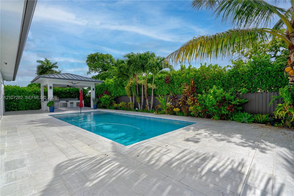In-ground swimming pool with stone-tiled decking and a covered outdoor kitchen featuring built-in cabinetry and professional-grade appliances, enclose