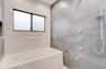 Jetted bathtub featuring textured tile surround, large-format grey wall tile, chrome shower fixtures with handheld sprayer, and rectangular window