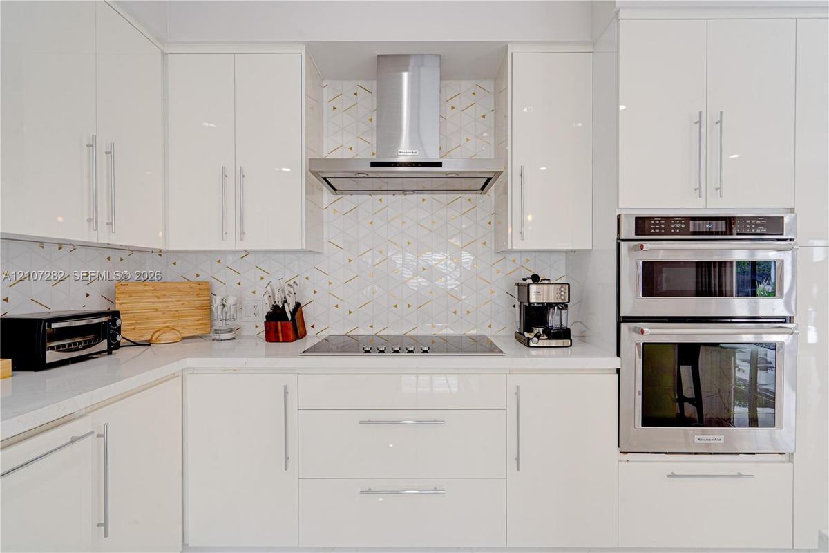 Kitchen featuring high-gloss white cabinetry, geometric patterned backsplash, stainless steel double wall oven, stainless steel range hood, and solid-