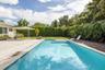 In-ground swimming pool with built-in steps, expansive wood-finish decking, light-colored stone patio surround, manicured lawn, and perimeter hedging