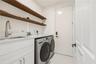 Laundry room featuring white cabinetry, stone-finish countertops, an undermount sink with a high-arch faucet, wall-mounted wood-finish shelving, and l