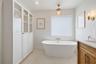 Freestanding soaking tub with wall-mounted fixtures, built-in floor-to-ceiling cabinetry with glass-panel doors, neutral tile flooring, wood-finish va