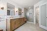 Dual-sink vanity with stone countertop, walk-in glass-enclosed shower featuring gold-tone hardware, wall-mounted light fixtures, and frosted glass doo