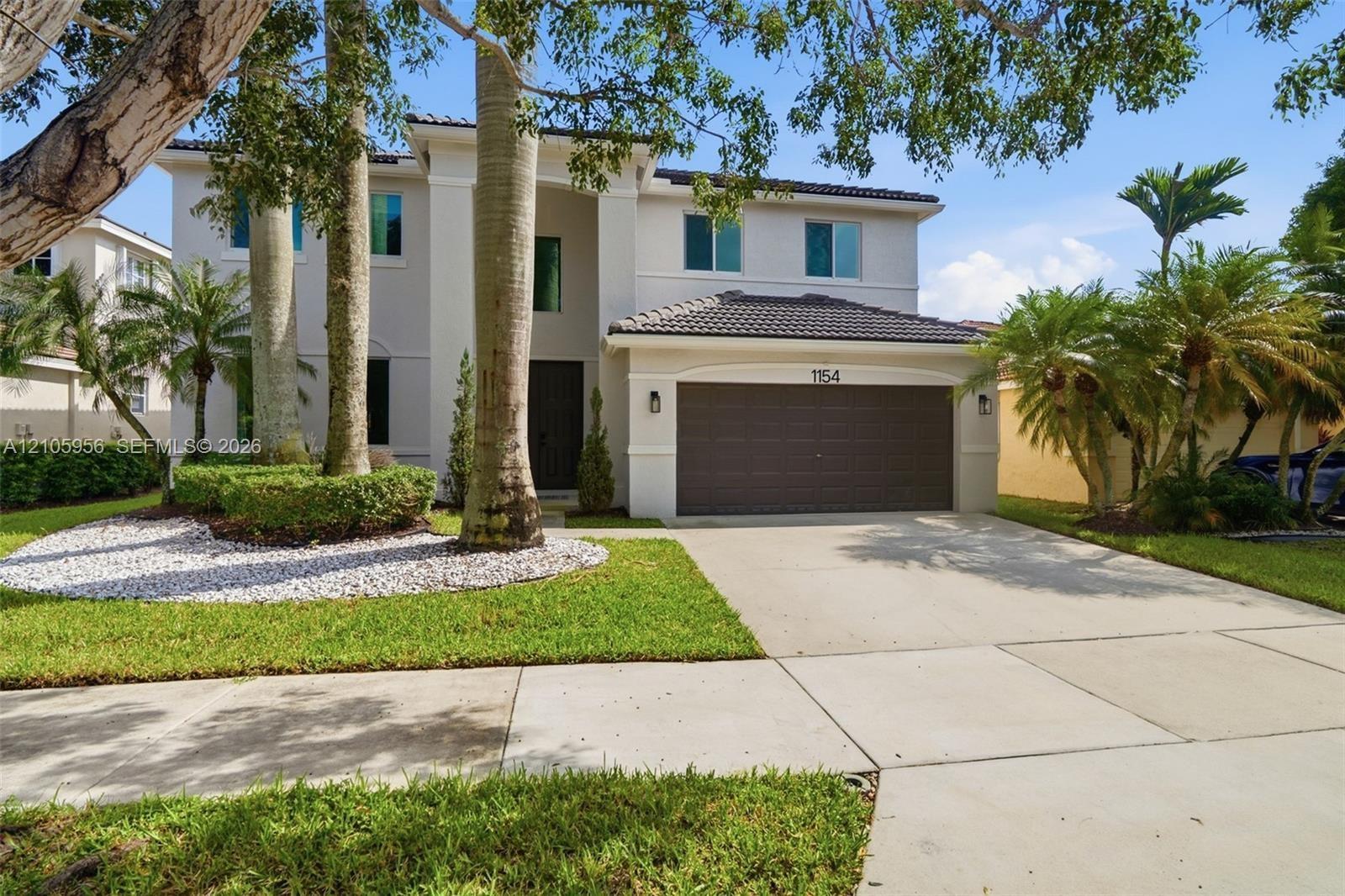 Two-story residence featuring a light-toned exterior, concrete tile roof, multi-car garage, and landscaped front grounds with mature palm trees