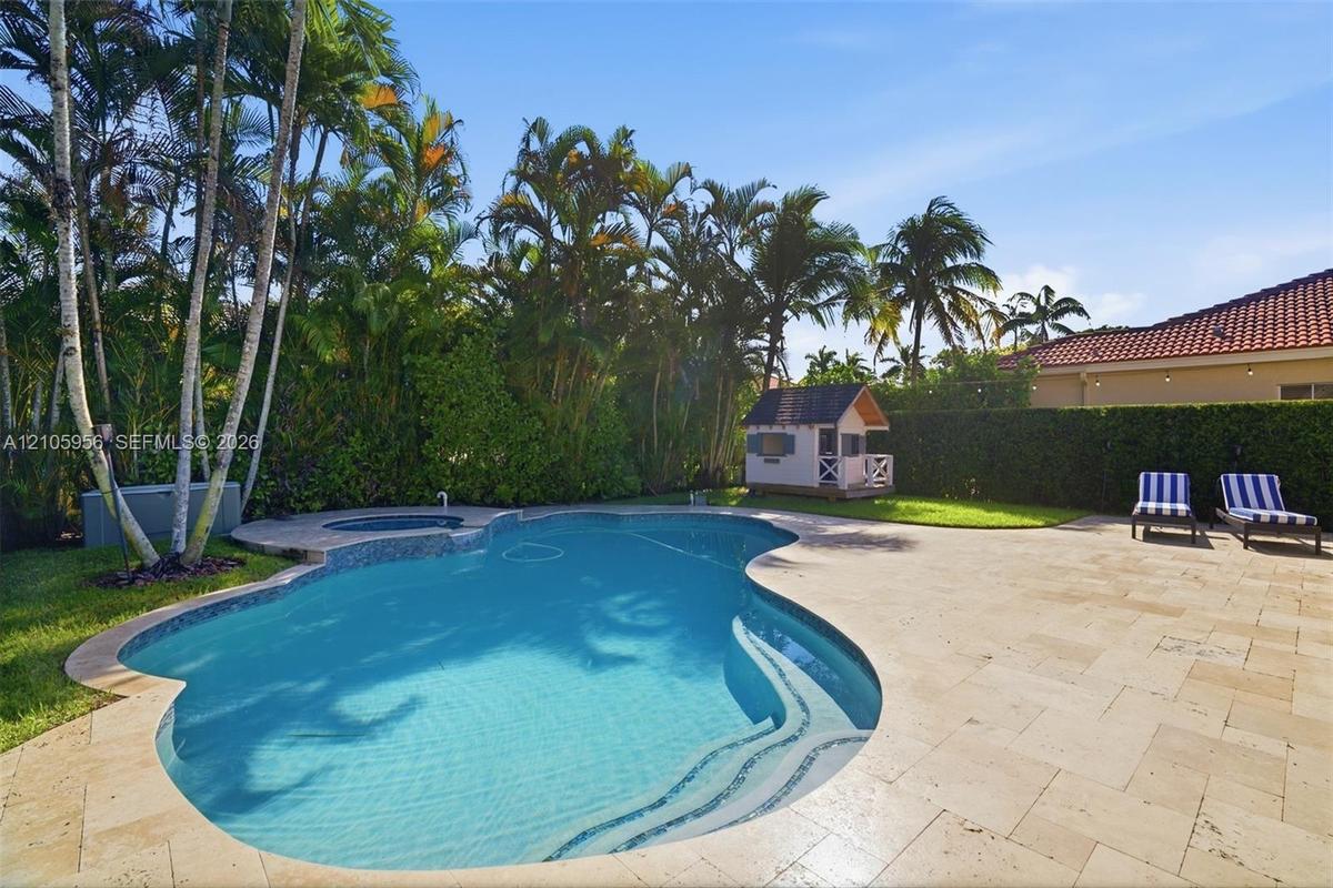 Free-form swimming pool featuring an integrated spillover spa, wide stone-paver decking, mature perimeter palm trees, and an enclosed lawn