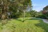 Expansive backyard featuring a manicured lawn, mature trees, dense perimeter landscaping, and a paved walkway