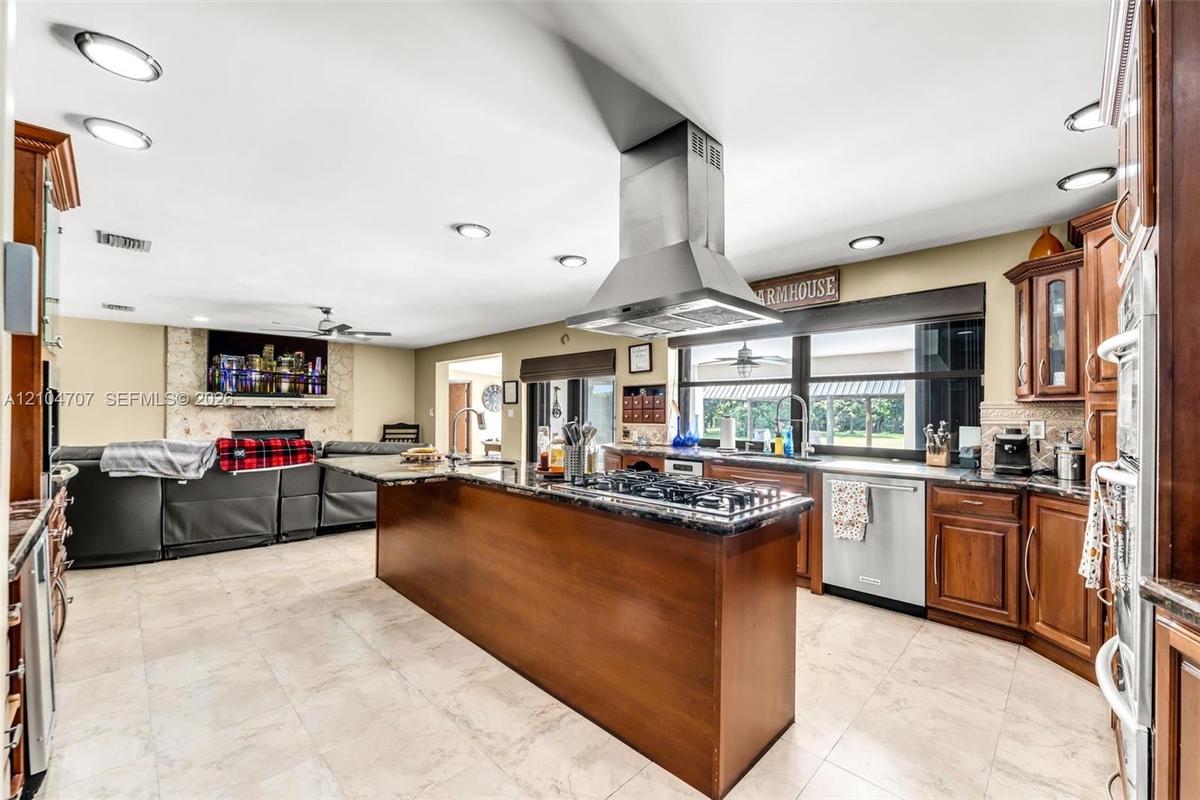 Kitchen featuring wooden cabinetry, a central island with an integrated gas cooktop, a stainless steel chimney-style range hood, stone-finish tile flo