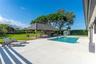 Backyard featuring a concrete pool deck, rectangular swimming pool, open-air pavilion with a metal roof, expansive grass lawn, and perimeter hedging