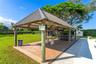 Expansive grass lawn featuring mature trees, perimeter hedge foliage, metal fencing, and a covered structure