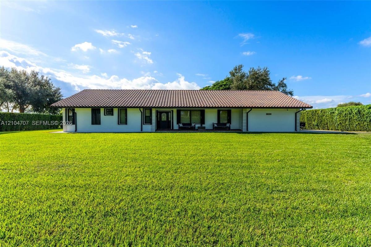 Single-story residence featuring a low-pitched tiled roof, light-toned exterior finish, and an expansive manicured lawn bordered by mature hedge fenci