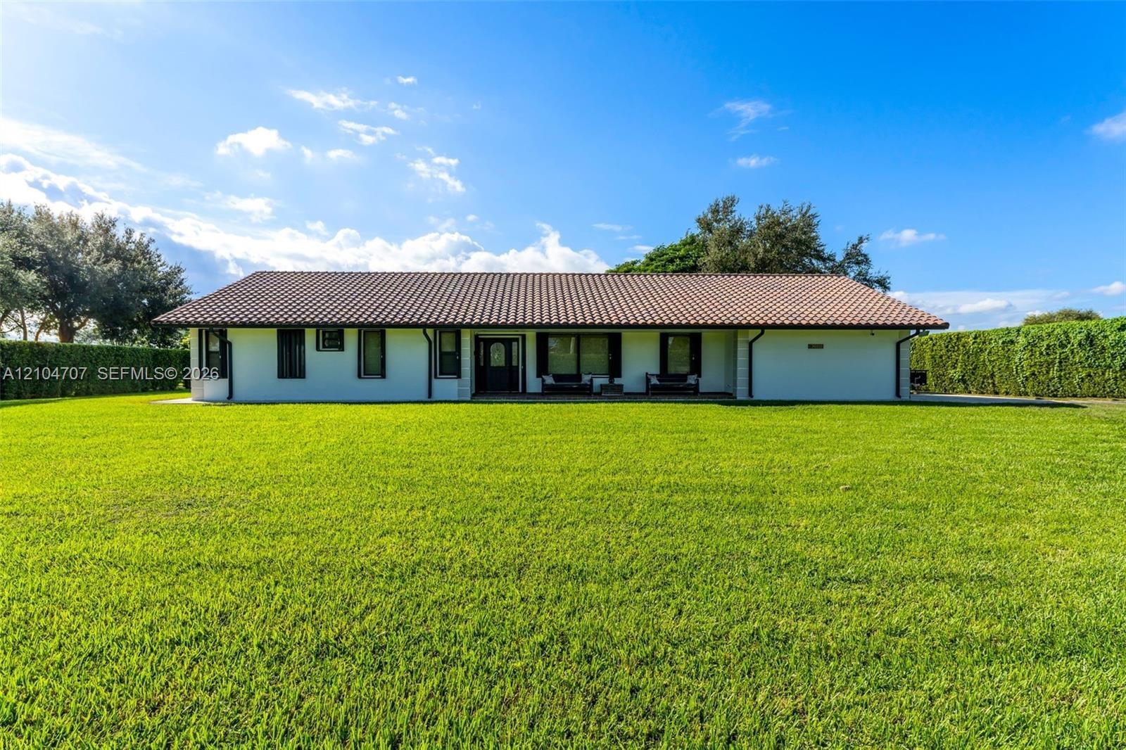 Single-story residence featuring a low-pitched tiled roof, light-toned exterior finish, and an expansive manicured lawn bordered by mature hedge fenci