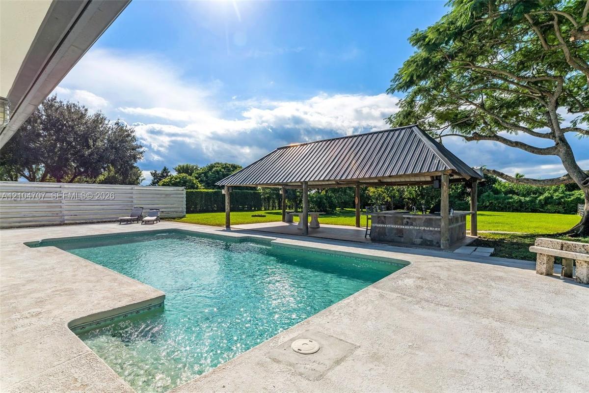 Outdoor living area featuring a swimming pool, covered pavilion with stone-finish bar, sprawling lawn, and mature canopy tree