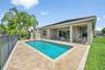 Covered patio with stone-paver flooring, rectangular swimming pool, and metal perimeter fencing overlooking a pond