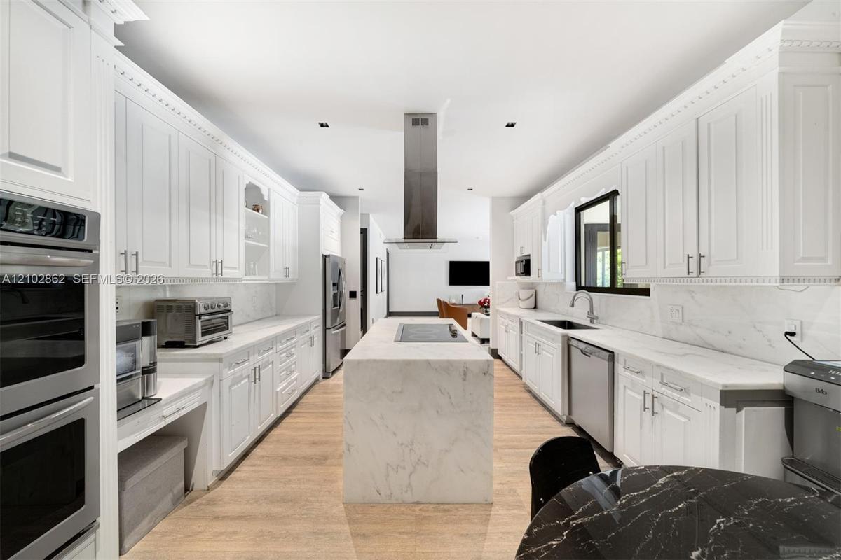 Gourmet kitchen featuring white shaker cabinetry, a waterfall-edge center island with integrated cooktop, stone-finish countertops, stainless steel wa