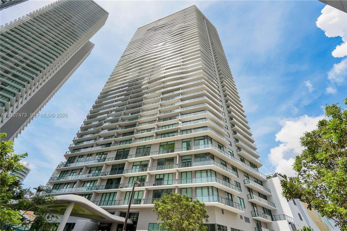 High-rise tower exterior featuring a multi-story facade, cantilevered balconies with glass railings, large-pane windows, and a structured concrete arc