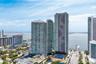 Aerial perspective of a high-rise residential complex featuring glass-facade towers, outdoor swimming pools, and tennis courts situated along the wate