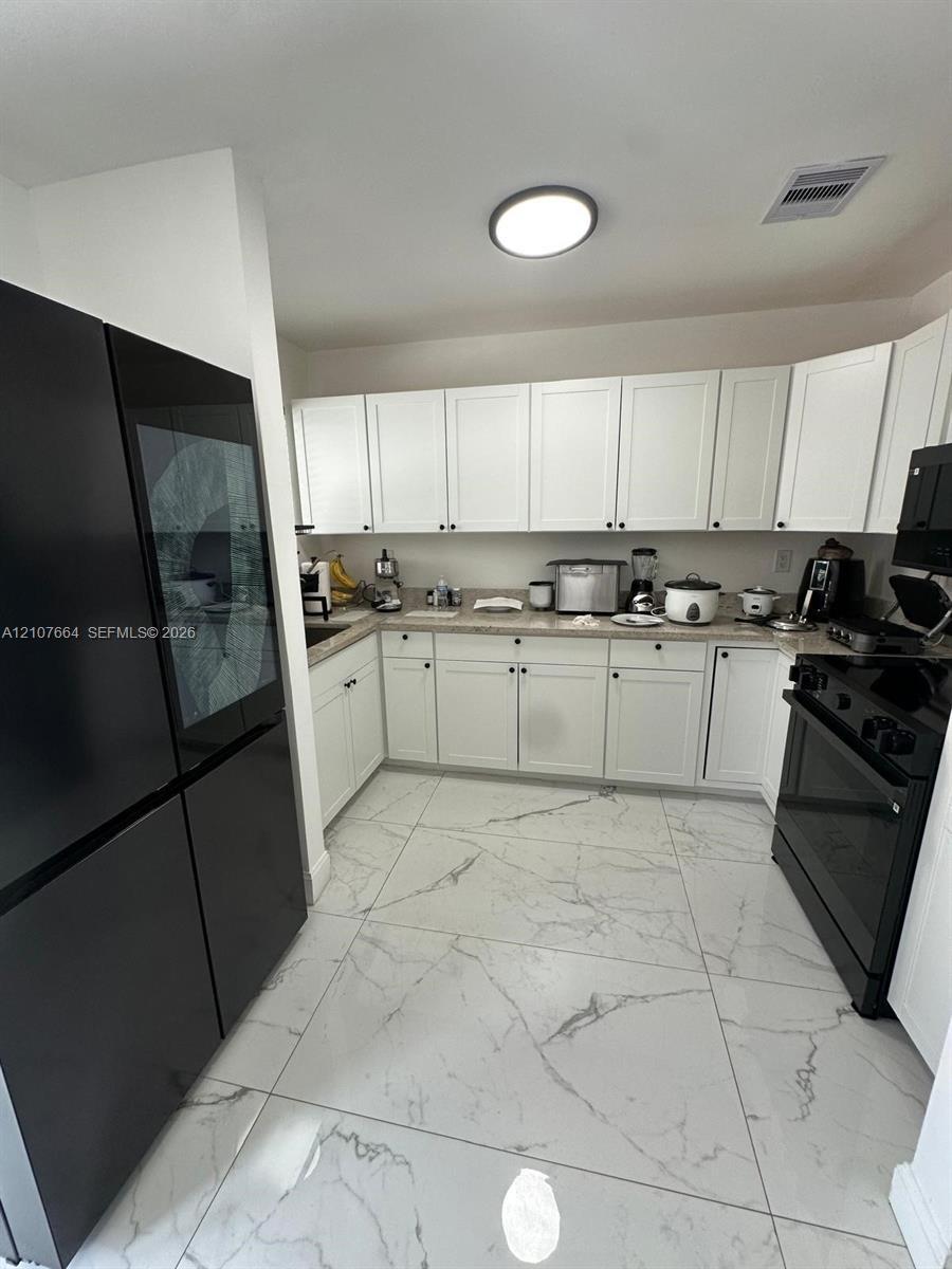 Kitchen featuring white shaker-style cabinetry, solid-surface countertops, large-format tile flooring with veined detailing, and black appliances