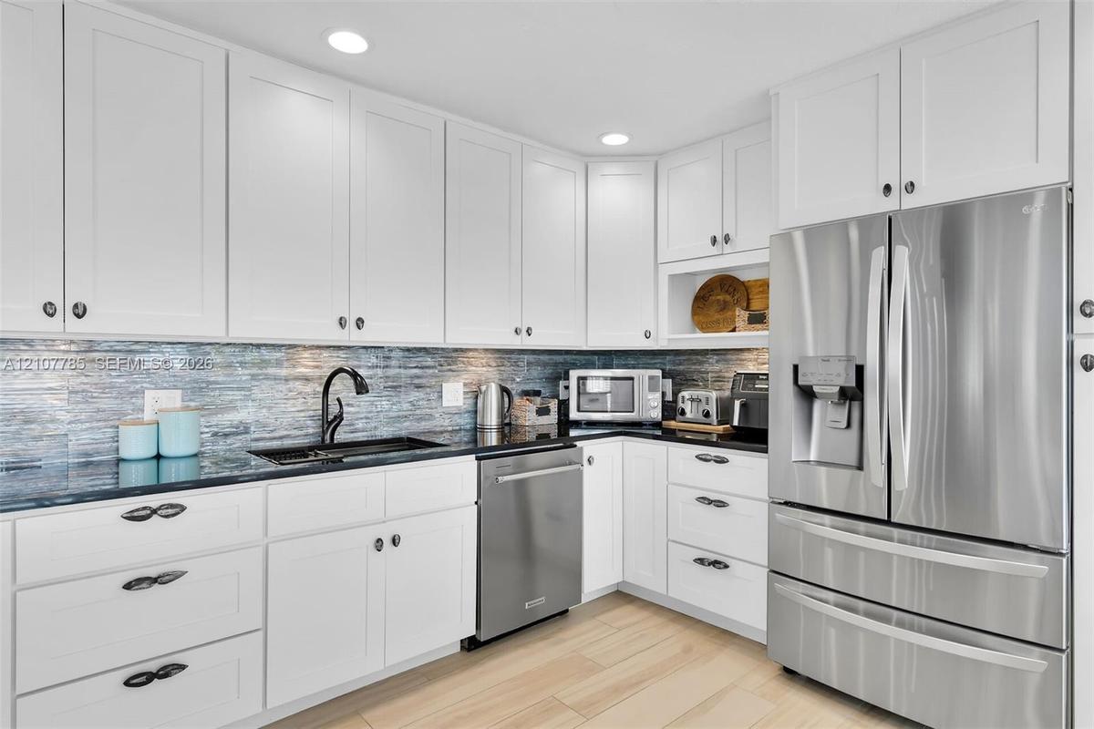 Kitchen featuring white shaker-style cabinetry, dark stone countertops, blue glass tile backsplash, stainless steel appliances, and wood-finish floori