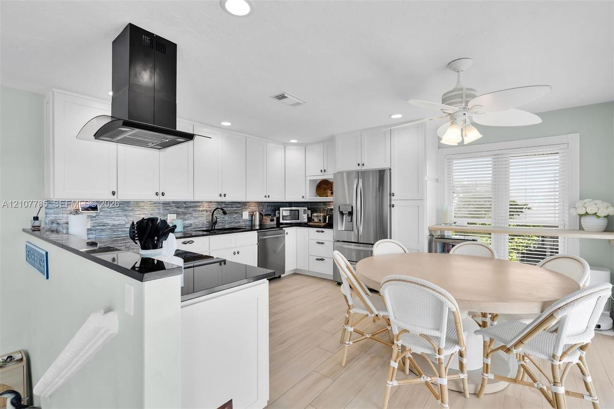 Kitchen featuring white shaker-style cabinetry, dark stone countertops, a stainless steel refrigerator, a black wall-mounted range hood, and wood-fini