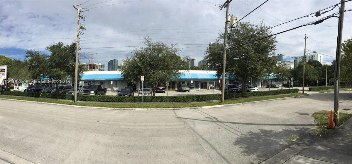 Single-story commercial plaza featuring an expansive surface parking lot, blue metal awnings, glass storefronts, and manicured perimeter hedging