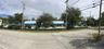 Single-story commercial plaza featuring an expansive surface parking lot, blue metal awnings, glass storefronts, and manicured perimeter hedging
