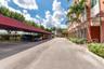 Asphalt parking area featuring covered vehicle shelters, multi-story building exterior with vertical windows, and manicured tropical landscaping with 