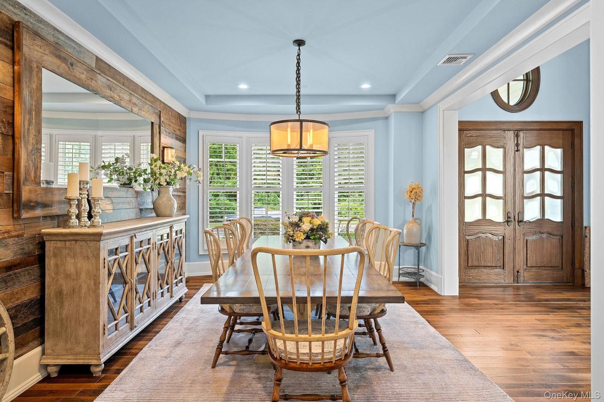 Dining area featuring wood-finish plank flooring, wood-paneled accent wall, tray ceiling with recessed lighting, bay windows with plantation shutters,