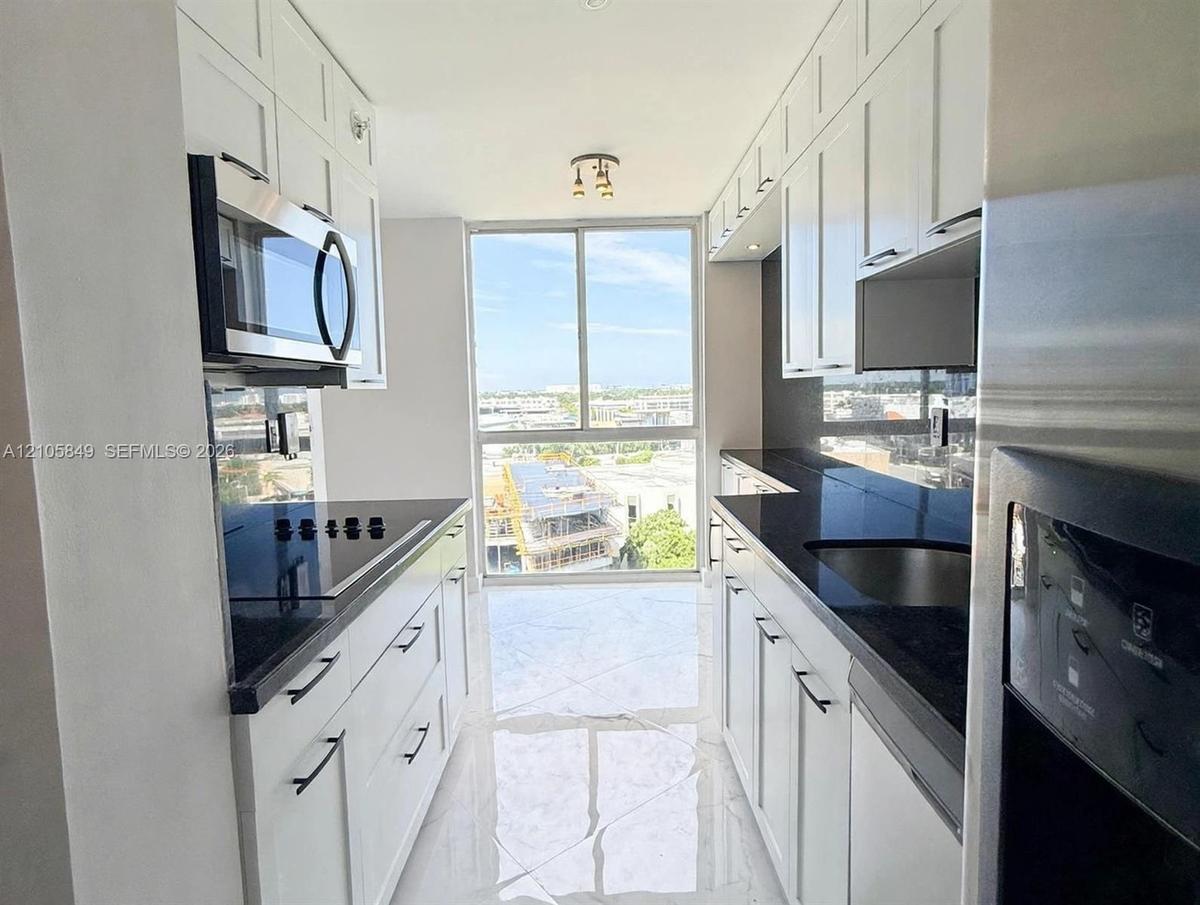 Galley kitchen featuring white Shaker-style cabinetry, dark stone countertops, stainless steel appliances, large-format tile flooring, and floor-to-ce