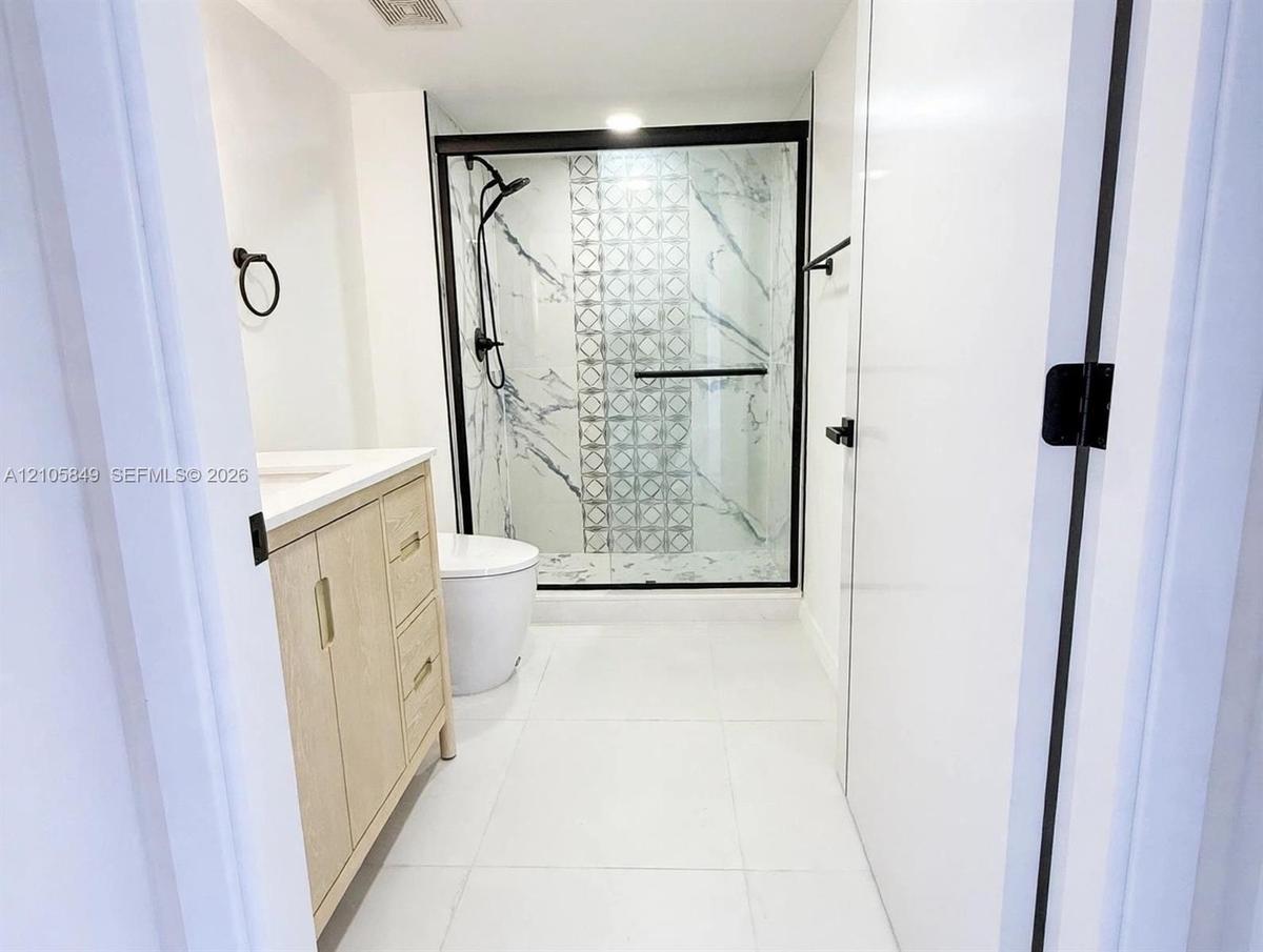 Bathroom featuring a light wood-finish vanity, large-format tile flooring, and a glass-enclosed shower with decorative accent tile, stone-patterned wa