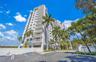 High-rise residential building featuring white stucco facade, stacked balconies, and paved surface parking, set amidst mature palm trees and manicured