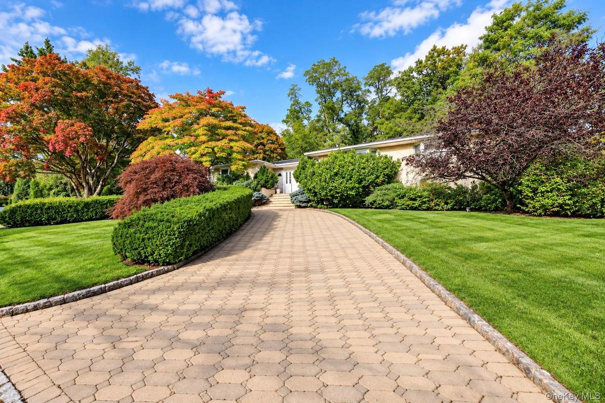 Stone paver driveway with Belgian block curbing, flanked by manicured lawns, shaped shrubbery, and mature deciduous trees