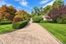 Stone paver driveway with Belgian block curbing, flanked by manicured lawns, shaped shrubbery, and mature deciduous trees