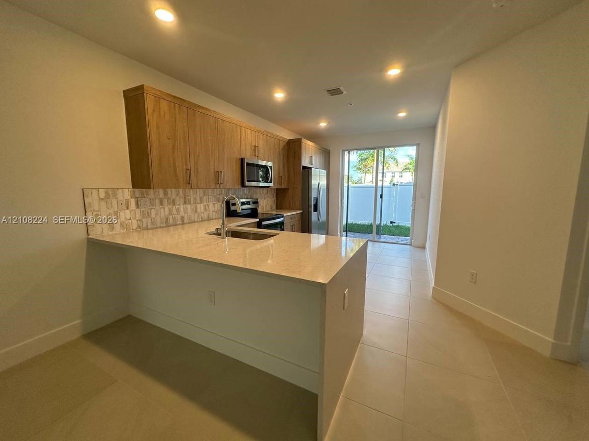 Kitchen featuring wood-finish cabinetry, quartz countertops, a stone-tile backsplash, stainless steel appliances, and large-format tile flooring
