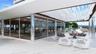 Covered outdoor lounge with large-format tile flooring and a decorative overhead canopy, featuring a glass-enclosed fitness center with high-gloss cyl