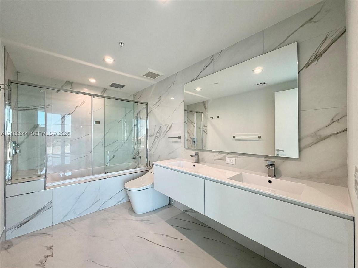 Floating double vanity with integrated sinks, full-wall mirror, glass-enclosed soaking tub with shower combination, and large-format porcelain tile wa