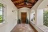 Covered entryway featuring terracotta tile flooring, exposed wood-beam vaulted ceiling, arched masonry openings, stone door surround with column detai