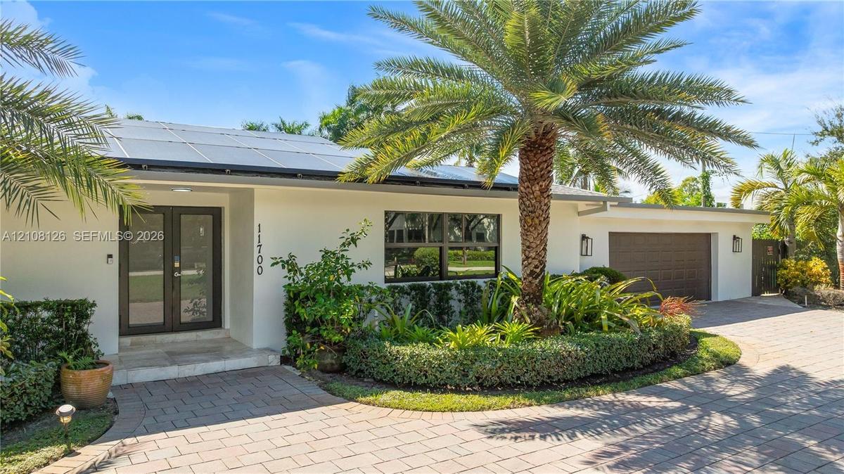 Single-story exterior featuring a light-toned stucco facade, rooftop solar panels, a paved brick driveway, a double-entry glass door, and integrated o