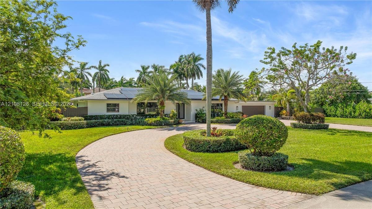 Single-story exterior featuring a paver circular driveway, solar panel installation, manicured landscaping with mature palm trees, and an attached two