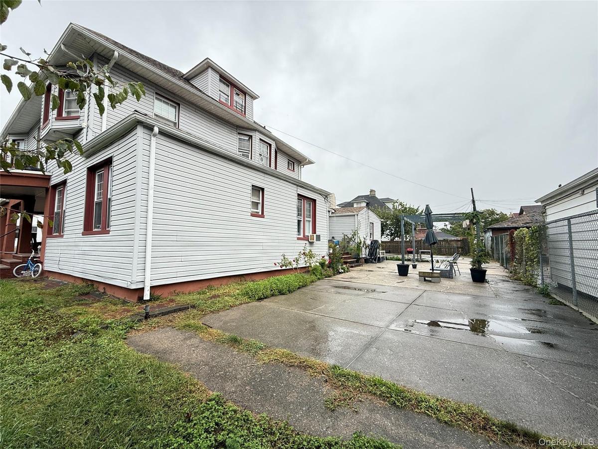 Exterior featuring light-colored horizontal siding, a multi-story roofline with dormers, a concrete patio area, and chain-link fencing