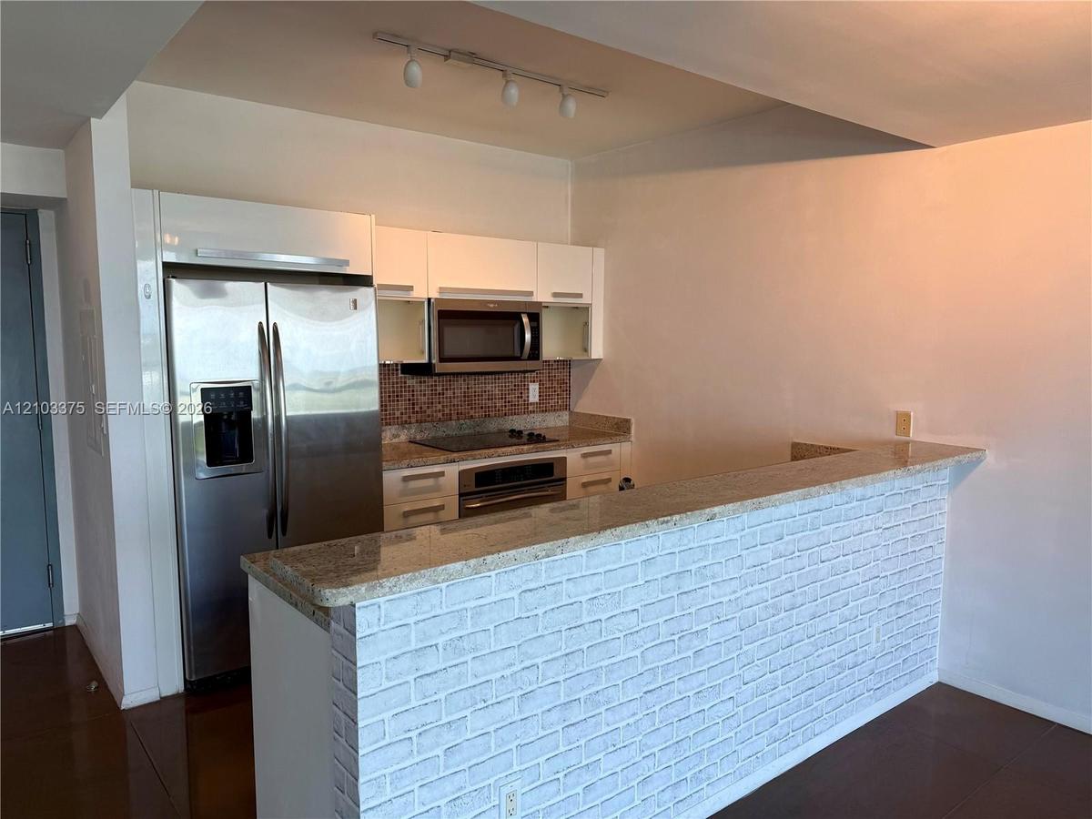 Kitchen featuring white flat-panel cabinetry, stainless steel appliances, stone-surface breakfast bar with a white brick base, and mosaic tile backspl